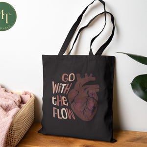 May include: Black tote bag with the words "GO WITH THE FLOW" and a maroon anatomical heart graphic. The bag has black handles and is made of a sturdy fabric. The bag is sitting on a wooden surface.