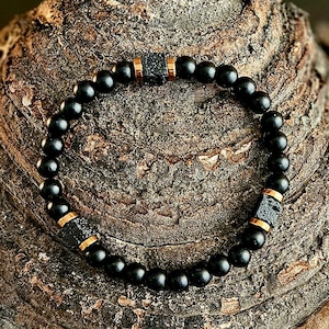 Könnte beinhalten: Ein schwarzes Perlenarmband mit goldfarbenen Akzenten. Das Armband besteht aus kleinen, runden schwarzen Perlen, die mit größeren, strukturierten schwarzen Perlen und goldfarbenen zylindrischen Abstandshaltern durchsetzt sind. Das Armband wird auf einer strukturierten, braunen Oberfläche präsentiert.