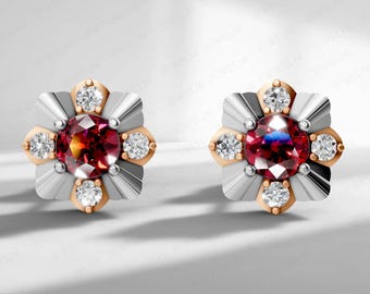 May include: A pair of stud earrings featuring a central, round, deep red gemstone surrounded by smaller, clear gemstones. The earrings have a silver and gold-tone flower design, with a light background.