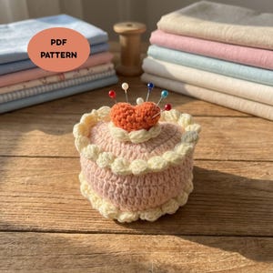 May include: A handmade crochet pin cushion in the shape of a cake, with a heart-shaped pin holder on top. The pin cushion is pink with cream-colored trim and has several pins with colorful heads. The image also includes stacks of fabric and the text "PDF PATTERN".