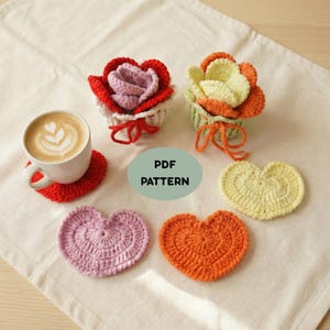 May include: A collection of handmade crochet items, including heart-shaped coasters in pink, orange, and yellow, and flower-shaped cup cozies in red, pink, and orange. A cup of coffee sits on a red coaster. The text "PDF PATTERN" is visible.