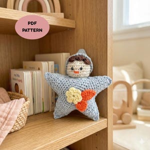May include: A handmade, light blue crochet star-shaped doll with a small doll head on top. The doll has brown hair, a beige face, and an orange mouth. It is decorated with yellow and orange flowers. The image also shows a bookshelf with books and a basket.