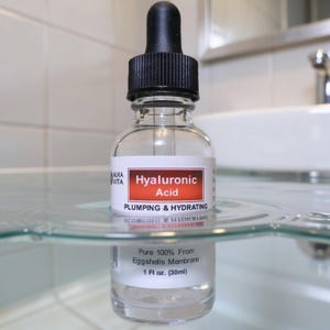 May include: A clear glass dropper bottle of Alka Vita Hyaluronic Acid serum with a black cap. The label reads Hyaluronic Acid, Plumping & Hydrating, and Pure 100% From Eggshells Membrane, 1 fl oz. The bottle is partially submerged in clear water.