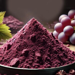 Puede incluir: Un mont&oacute;n c&oacute;nico de polvo de uva de color morado intenso se encuentra en un cuenco oscuro. El polvo fino y texturizado presenta peque&ntilde;os grumos. A un lado, un racimo de uvas moradas frescas y redondas y una hoja verde son visibles.