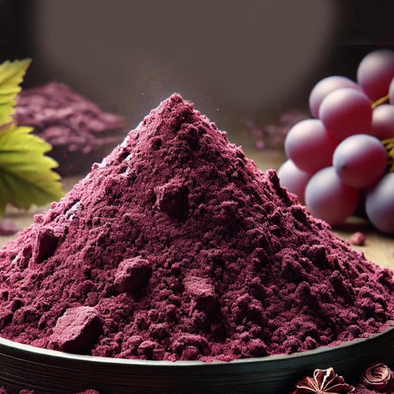 Puede incluir: Un mont&oacute;n c&oacute;nico de polvo de uva de color morado intenso se encuentra en un cuenco oscuro. El polvo fino y texturizado presenta peque&ntilde;os grumos. A un lado, un racimo de uvas moradas frescas y redondas y una hoja verde son visibles.