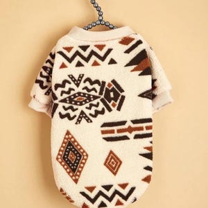 May include: A cream-colored dog sweater with a geometric pattern in brown and black. The sweater has a crew neck and short sleeves. The design includes zigzags, diamonds, and other shapes. The sweater is hanging on a beaded hanger.