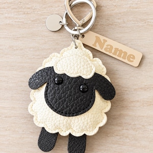 May include: A sheep-shaped keychain featuring a textured cream-colored body and a black face with two small black eyes. The keychain includes a silver-toned metal ring, a small circular charm, and a rectangular tag with the word Name printed on it.