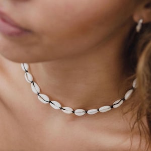 Pode incluir: Um colar de búzios brancos com fio preto. As conchas ovais são unidas, criando um estilo gargantilha. Um acessório natural e praiano.