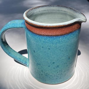 May include: A ceramic pitcher with a glossy turquoise blue glaze featuring a speckled finish. The rim is accented with a wide, textured band of reddish-brown clay. It includes a curved handle on the side and a small pouring spout at the top edge.