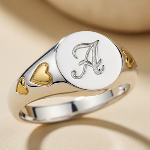 以下が含まれることがあります： 筆記体の「A」の文字が特徴の、白い円形の顔を持つシルバーリング。リングの両側には金のハートのアクセントが付いています。シグネットスタイルのリングです。
