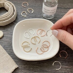 Puede incluir: Una colección de pequeños aros metálicos en forma de C en tonos plata, oro y oro rosa. Los aros se muestran dentro de un pequeño plato de cerámica blanca y en una botella de vidrio transparente con la etiqueta Stalking Hoops Mixed Metals.