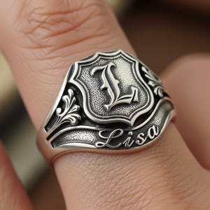 Puede incluir: Un anillo de sello plateado con un diseño de escudo ornamentado. El escudo presenta la letra "L" en una fuente estilizada. El nombre "Lisa" está grabado en la banda del anillo. El anillo se lleva en un dedo.