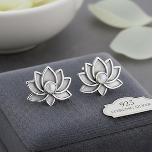 Op de afbeelding: Een paar zilveren lotusbloem oorknopjes. Elke oorbel heeft een gedetailleerd lotusontwerp met een centrale parel. De oorbellen worden gepresenteerd op een grijze fluwelen ondergrond, met een label "925 STERLING SILVER".