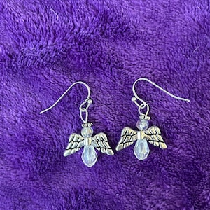 May include: A pair of silver-toned angel earrings with clear crystal-like teardrop-shaped bodies and detailed wings. The earrings hang from silver-colored fish hook ear wires. The background is a textured, deep purple fabric.