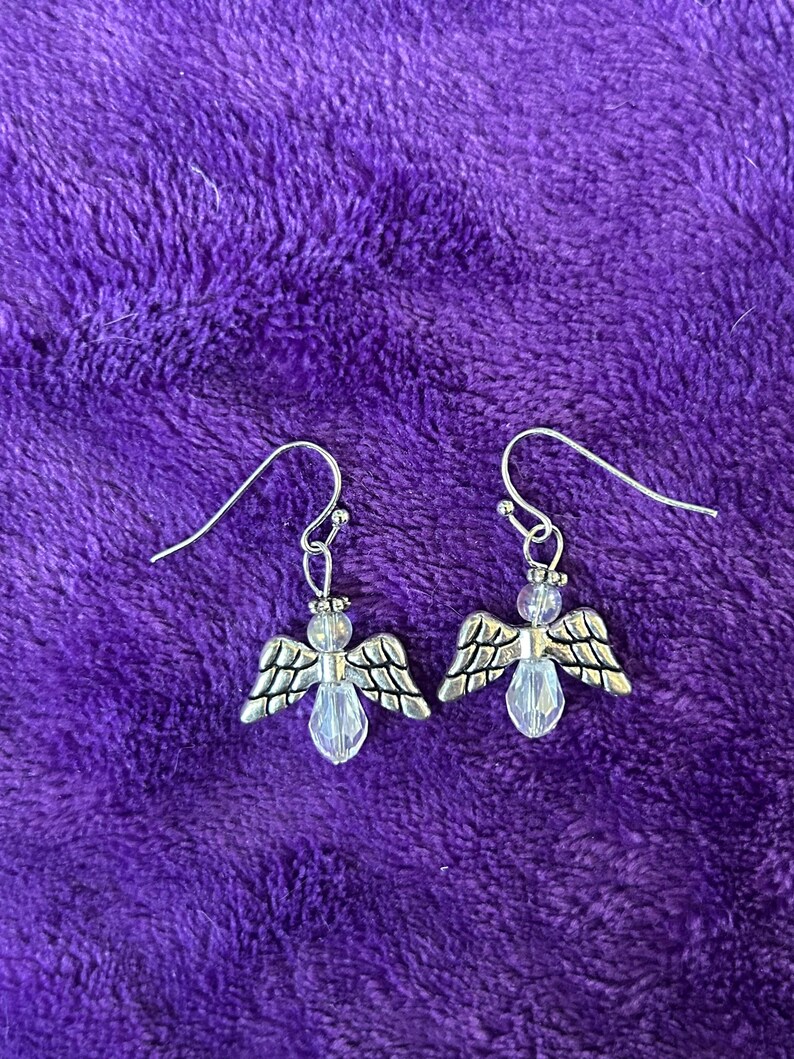 May include: A pair of silver-toned angel earrings with clear crystal-like teardrop-shaped bodies and detailed wings. The earrings hang from silver-colored fish hook ear wires. The background is a textured, deep purple fabric.