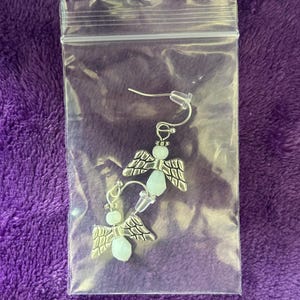 May include: A pair of angel-wing earrings in a clear plastic bag. The earrings have silver-colored wings and light blue beads. The earrings are on a purple background.