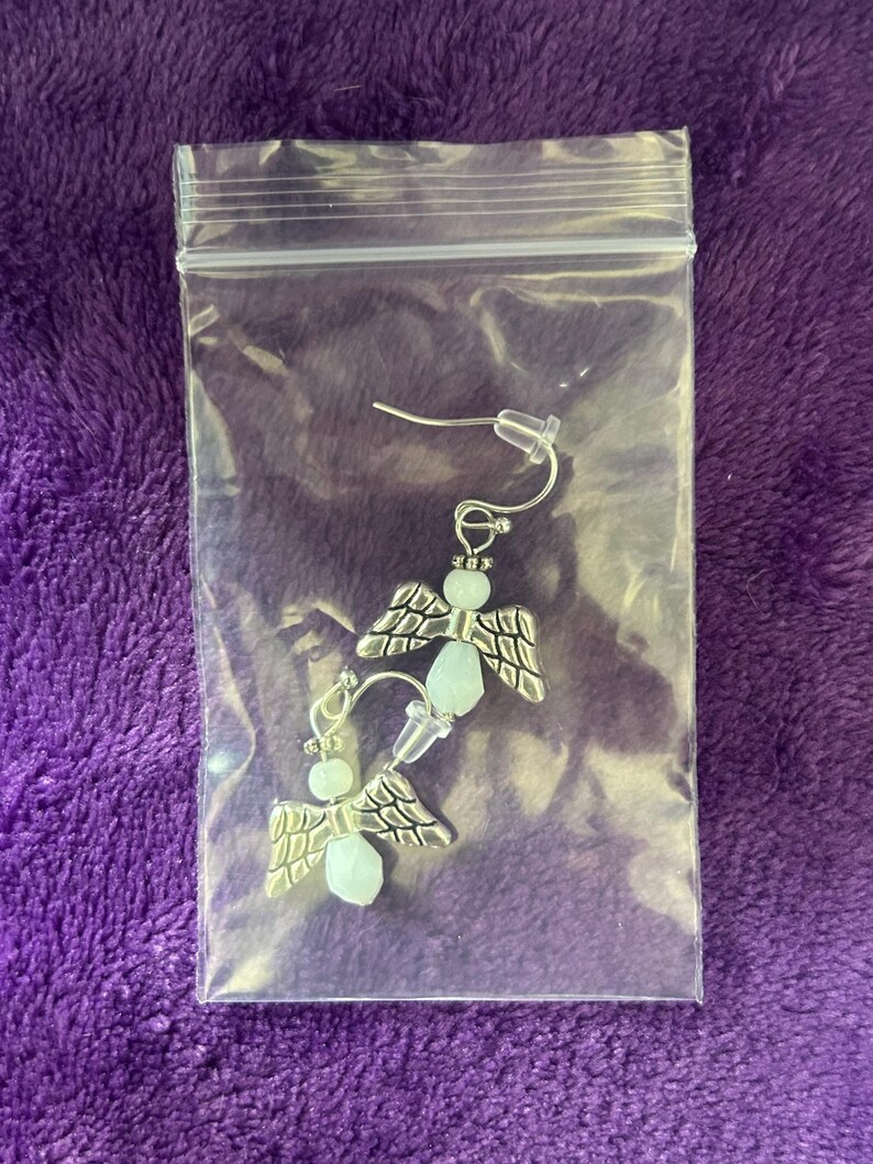May include: A pair of angel-wing earrings in a clear plastic bag. The earrings have silver-colored wings and light blue beads. The earrings are on a purple background.