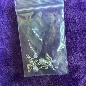 May include: A pair of silver-toned angel wing earrings with clear crystal accents, displayed in a clear plastic bag. The earrings have a detailed wing design and are on a purple background.