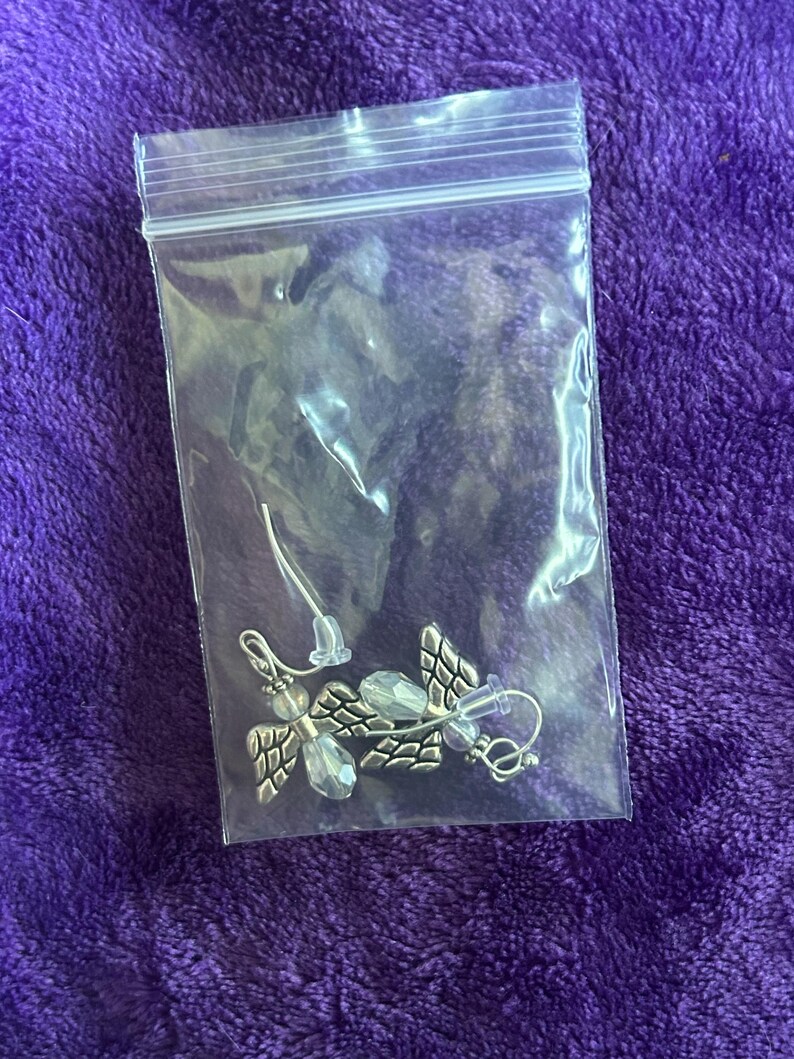 May include: A pair of silver-toned angel wing earrings with clear crystal accents, displayed in a clear plastic bag. The earrings have a detailed wing design and are on a purple background.