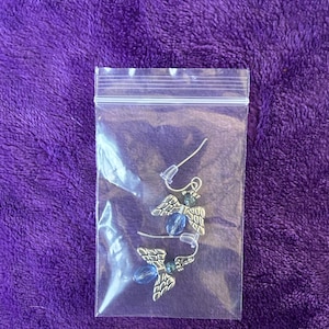 May include: A pair of silver-toned angel wing earrings with blue bead accents, packaged in a clear plastic bag. The earrings feature detailed wing designs and are displayed against a purple background.