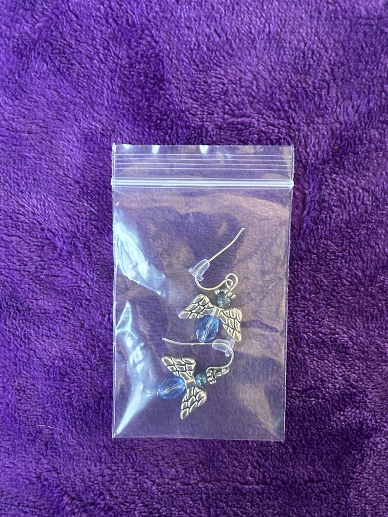 May include: A pair of silver-toned angel wing earrings with blue bead accents, packaged in a clear plastic bag. The earrings feature detailed wing designs and are displayed against a purple background.
