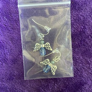 May include: A pair of angel-wing earrings in a clear plastic bag. The earrings have silver-colored wings, blue beads, and small green beads. The background is a textured purple fabric.