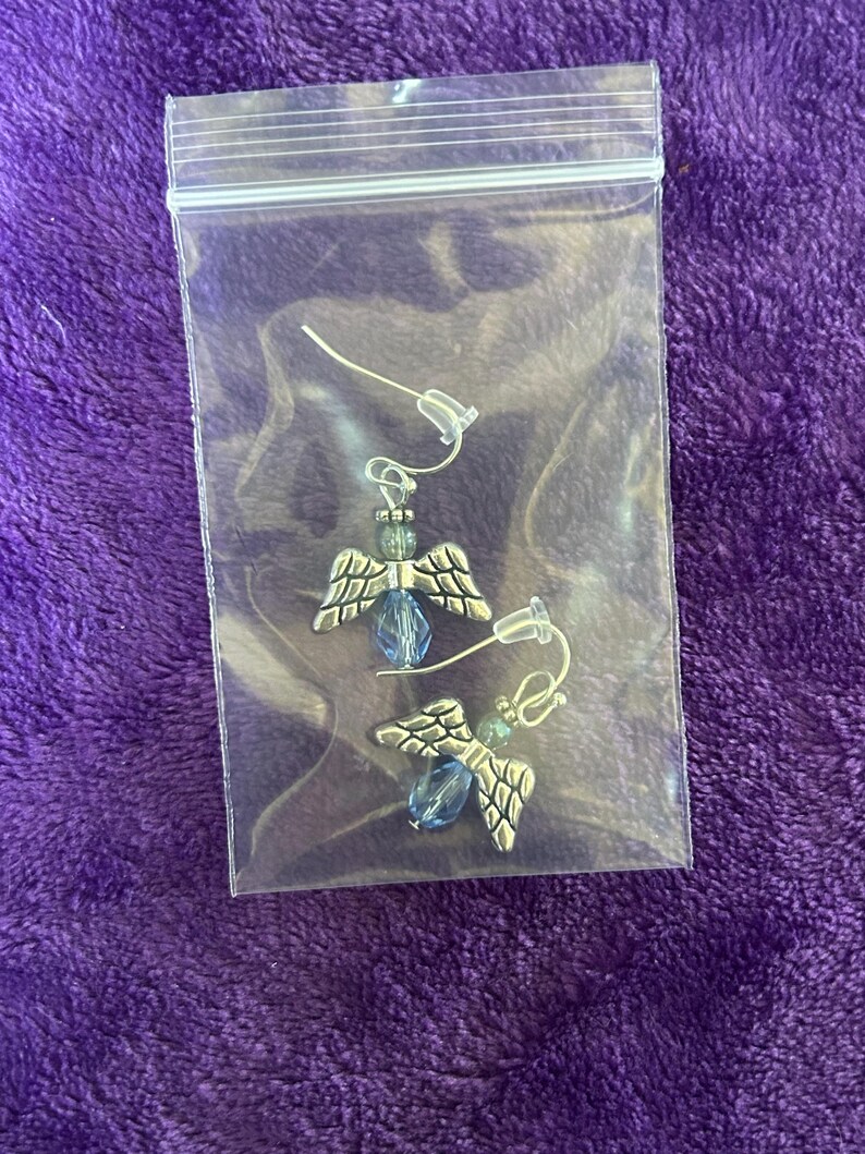 May include: A pair of angel-wing earrings in a clear plastic bag. The earrings have silver-colored wings, blue beads, and small green beads. The background is a textured purple fabric.