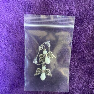 May include: A pair of angel earrings inside a clear plastic bag. The earrings feature silver-toned wings and white, oval-shaped beads for the body. The earrings are displayed on a purple, textured background.
