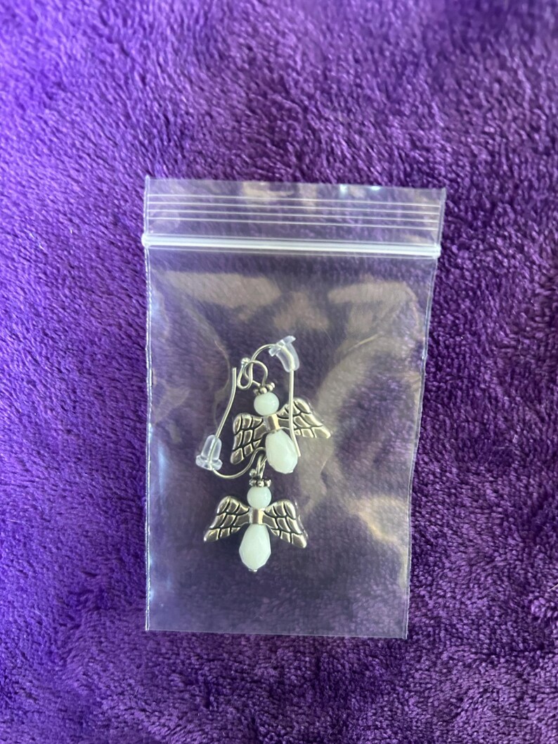 May include: A pair of angel earrings inside a clear plastic bag. The earrings feature silver-toned wings and white, oval-shaped beads for the body. The earrings are displayed on a purple, textured background.