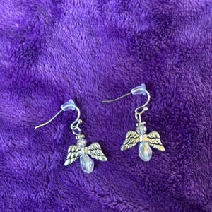 May include: A pair of silver-toned angel earrings with clear, teardrop-shaped crystal bodies and detailed wings. The earrings hang from silver-colored fish hook ear wires, set against a plush purple background.