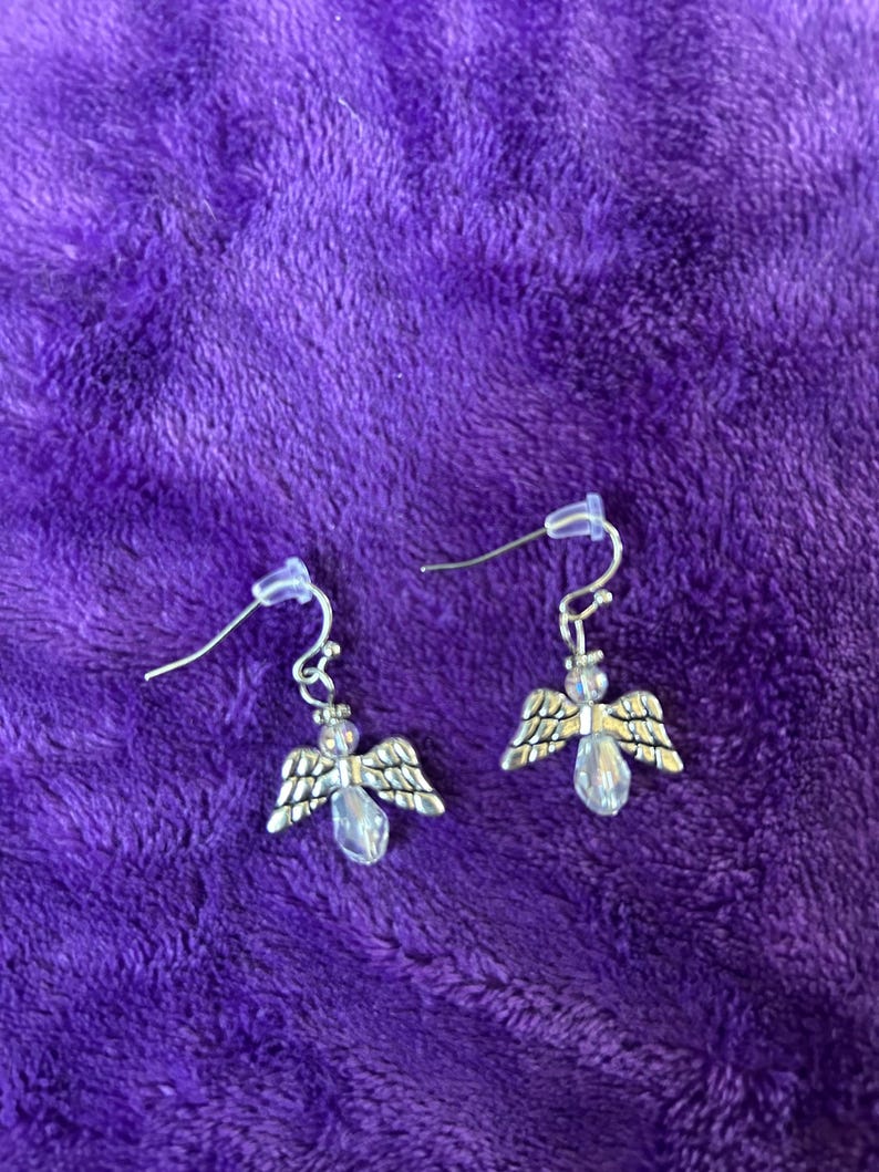 May include: A pair of silver-toned angel earrings with clear, teardrop-shaped crystal bodies and detailed wings. The earrings hang from silver-colored fish hook ear wires, set against a plush purple background.