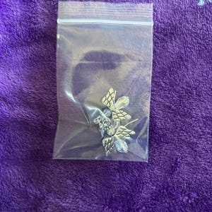 May include: A pair of silver-toned butterfly earrings with clear crystal accents, packaged in a clear plastic bag. The earrings have detailed wing designs and are displayed on a purple, textured surface.