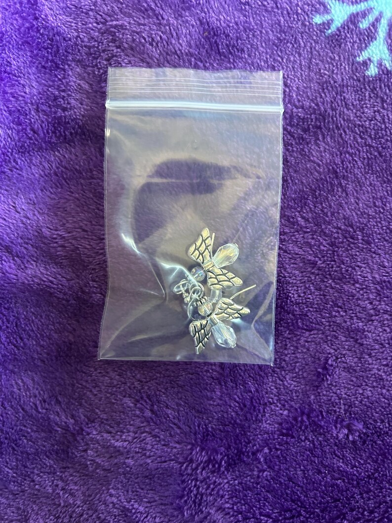 May include: A pair of silver-toned butterfly earrings with clear crystal accents, packaged in a clear plastic bag. The earrings have detailed wing designs and are displayed on a purple, textured surface.