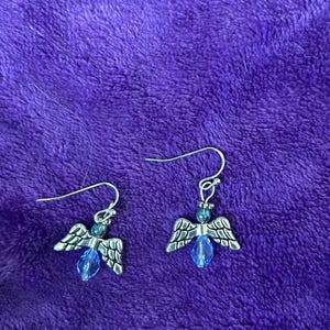 May include: A pair of silver-toned angel wing earrings with blue crystal accents. The earrings have a hook closure and are displayed on a purple, textured surface. The wings are detailed with etched patterns, and the crystals are teardrop-shaped.