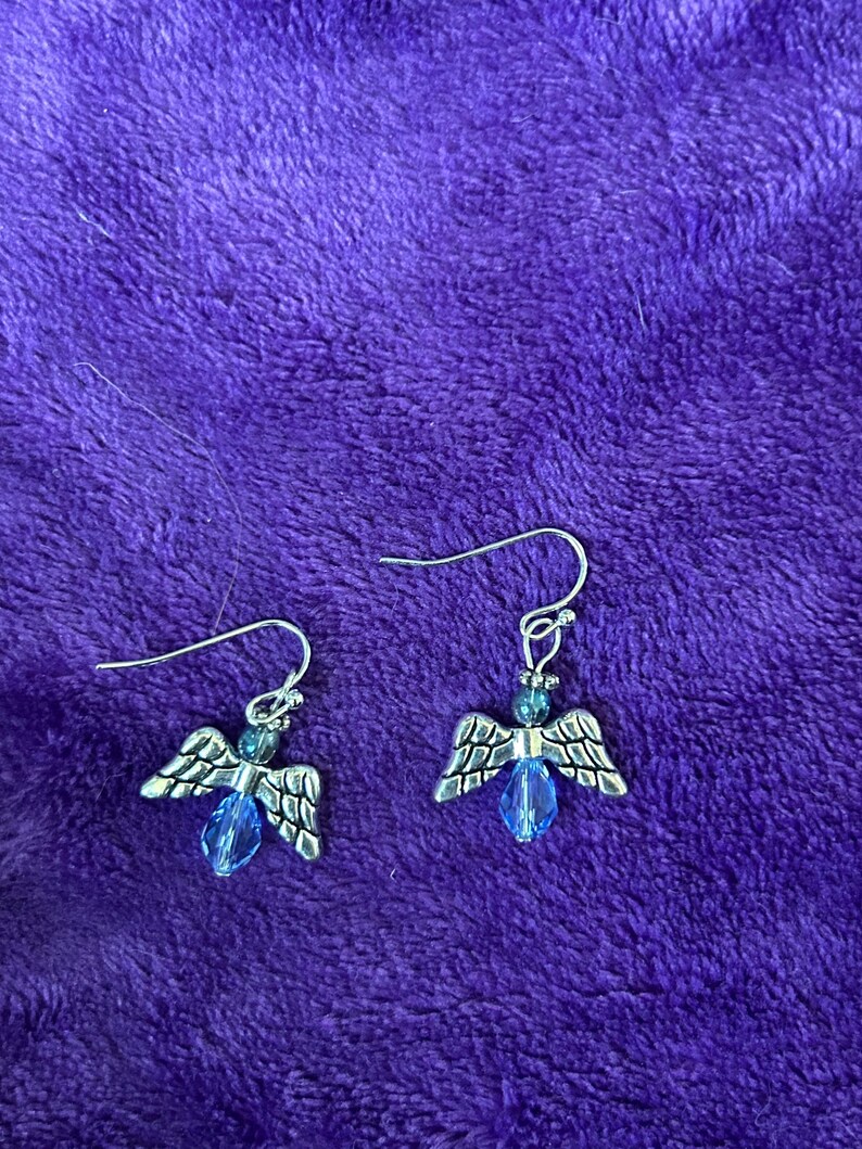 May include: A pair of silver-toned angel wing earrings with blue crystal accents. The earrings have a hook closure and are displayed on a purple, textured surface. The wings are detailed with etched patterns, and the crystals are teardrop-shaped.
