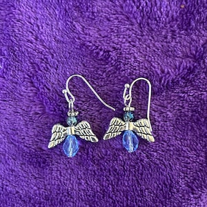 May include: A pair of silver-toned angel earrings with blue accents. Each earring features a small angel charm with detailed wings, a blue bead, and a silver hook. The earrings are set against a textured purple background.