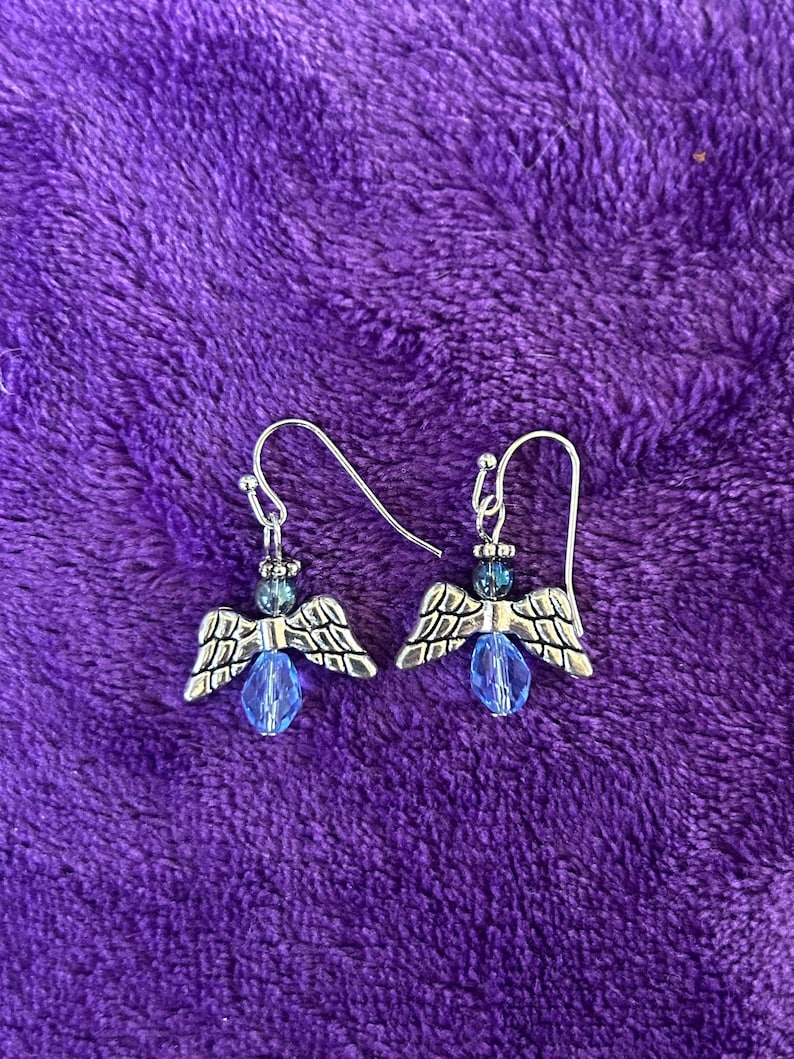 May include: A pair of silver-toned angel earrings with blue accents. Each earring features a small angel charm with detailed wings, a blue bead, and a silver hook. The earrings are set against a textured purple background.