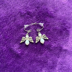 May include: A pair of silver-toned angel earrings with white bead accents and detailed wings. The earrings are displayed on a textured purple surface, showcasing their delicate design and craftsmanship.