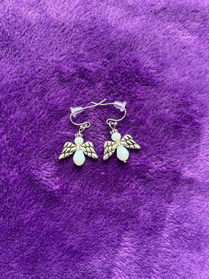 May include: A pair of silver-toned angel earrings with white bead accents and detailed wings. The earrings are displayed on a textured purple surface, showcasing their delicate design and craftsmanship.