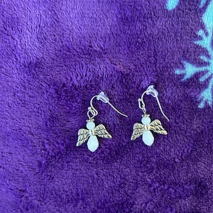 May include: A pair of silver-toned angel earrings. Each earring features a small angel charm with detailed wings and a pear-shaped, light-colored bead for the body. The earrings have a simple hook closure.