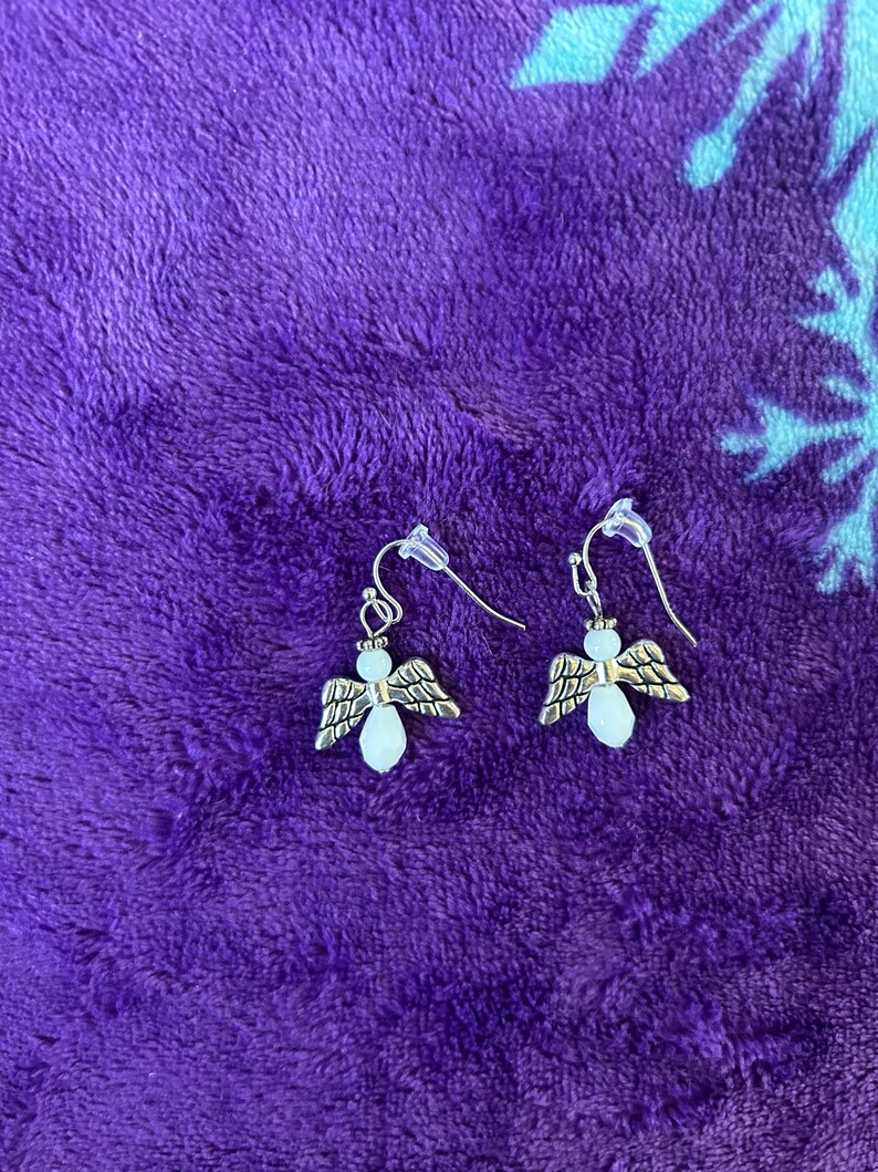 May include: A pair of silver-toned angel earrings. Each earring features a small angel charm with detailed wings and a pear-shaped, light-colored bead for the body. The earrings have a simple hook closure.