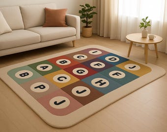 May include: A rectangular area rug with a colorful, block-style design featuring the letters A through J in white circles. The rug has a cream-colored border and is placed on a light-colored wooden floor. A beige sofa and a small wooden table are in the background.