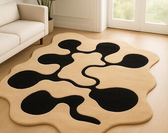 May include: A beige and black area rug with an abstract design. The rug features a beige background with a black, amoeba-like pattern. The rug is placed on a light wood floor in a room with a white sofa and a large plant.