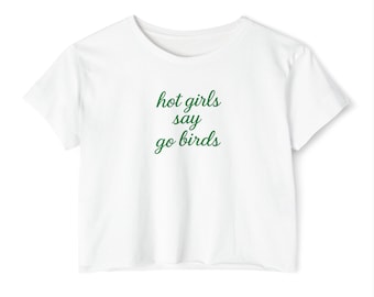Może przedstawiać: Biały top z napisem "hot girls say go birds" w zielonym kolorze. Koszulka ma okrągły dekolt i krótkie rękawy. Materiał wydaje się być miękki i lekki.