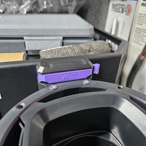 Può includere: Un componente di altoparlante audio nero e viola con le lettere "BC" visibili. L'altoparlante è montato su un altoparlante nero più grande. Lo sfondo include strumenti e contenitori di stoccaggio.