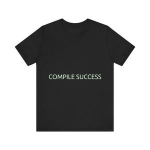 May include: Black t-shirt with the text "COMPILE SUCCESS" in a light green font. The short-sleeved shirt is a solid black color and has a crew neck.