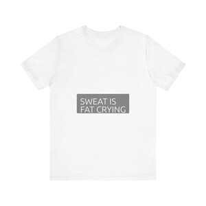 Może przedstawiać: Biała koszulka z napisem "SWEAT IS FAT CRYING" w szarym prostokącie. Koszulka ma okrągły dekolt i nadaje się do noszenia na co dzień lub na treningi. Tekst jest prostą czcionką bezszeryfową.