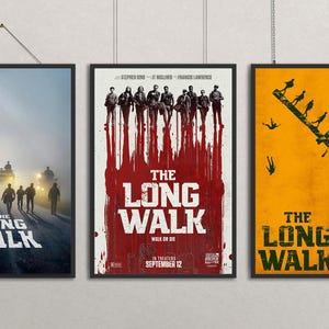 Puede incluir: Se muestran tres carteles enmarcados de la película "The Long Walk". Los carteles presentan diferentes diseños, incluyendo un grupo de personas caminando por una carretera, un fondo rojo sangre con figuras y un fondo amarillo con siluetas.
