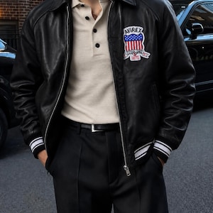 May include: A black leather bomber jacket with a silver zipper, a pointed collar, and striped ribbed cuffs. A patch on the left chest features the text AVIREX, a shield with red and white stripes and blue stars, and the text ESTABLISHED 1975.