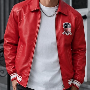 May include: A bright red leather bomber jacket featuring a silver front zipper, a classic collar, and ribbed cuffs with white stripes. An embroidered patch on the left chest displays the text AVIREX above a shield with a flag design and 1975.